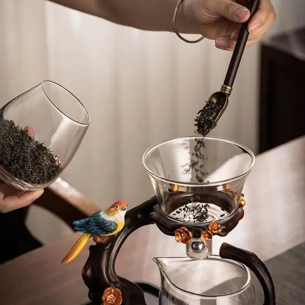 Person adding tea leaves to a glass teapot with a decorative bird handle.