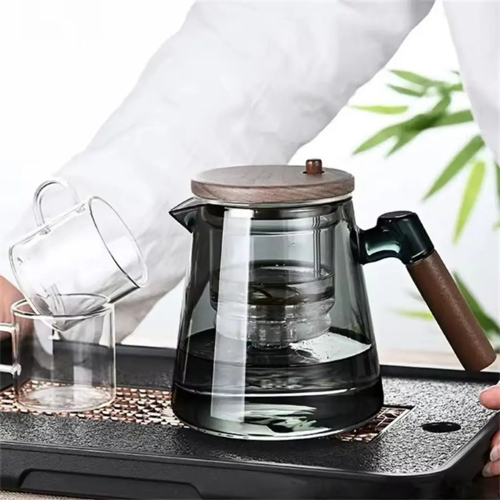 Glass teapot with wooden lid and handle on a black tray, person pouring from clear glass mug.