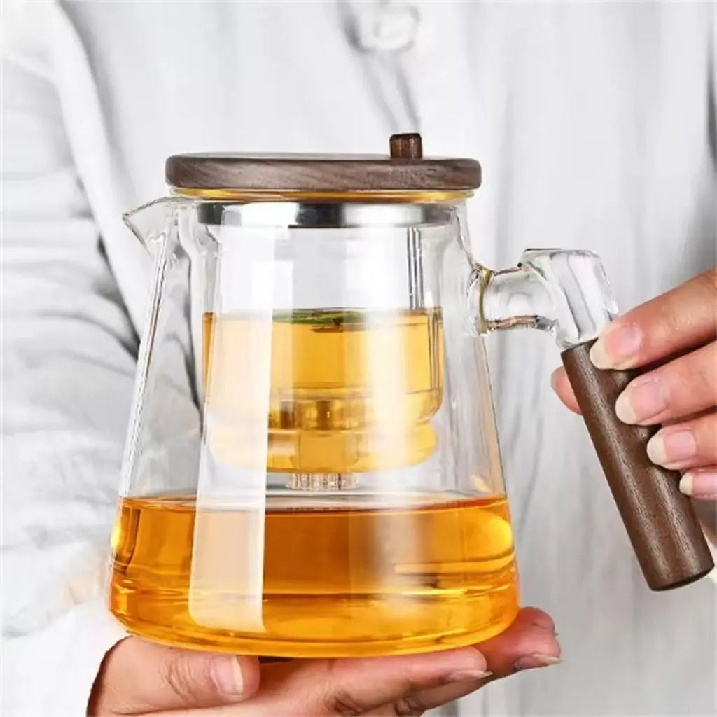 Clear glass teapot with wooden lid and handle filled with tea, held by a person.