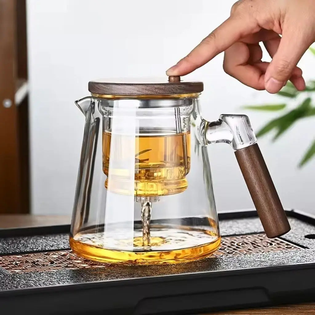 Glass teapot with wooden handle and lid being filled with tea, on a table.