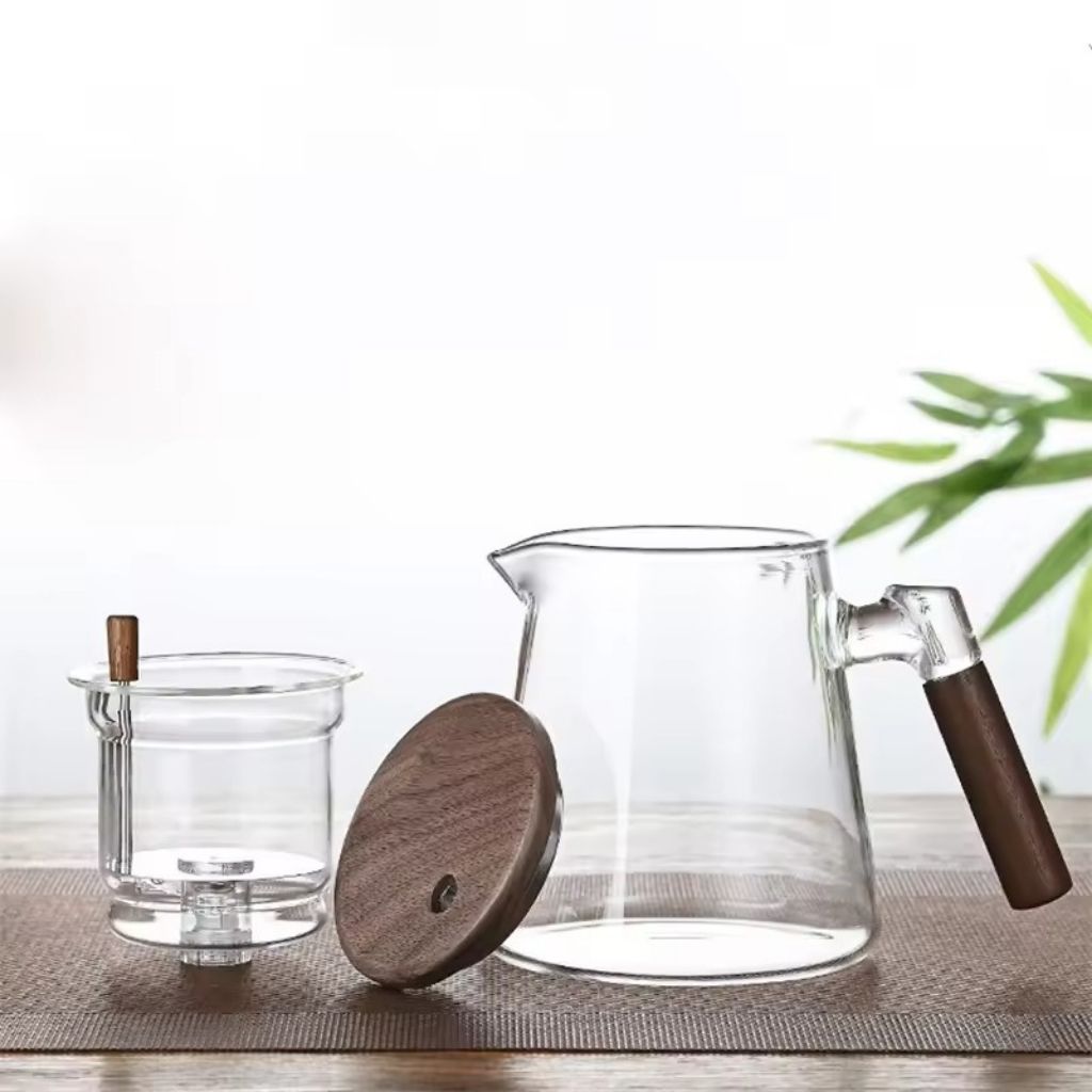 Clear glass pitcher with wooden handle and lid on a light surface with a blurred green plant in the background