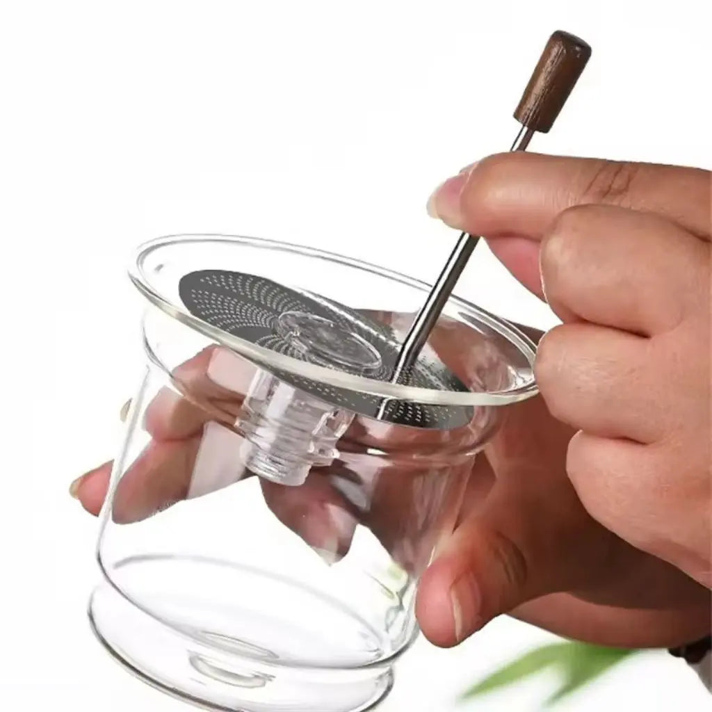 Clear glass tea infuser with metal mesh and wooden handle held by a hand against a white background