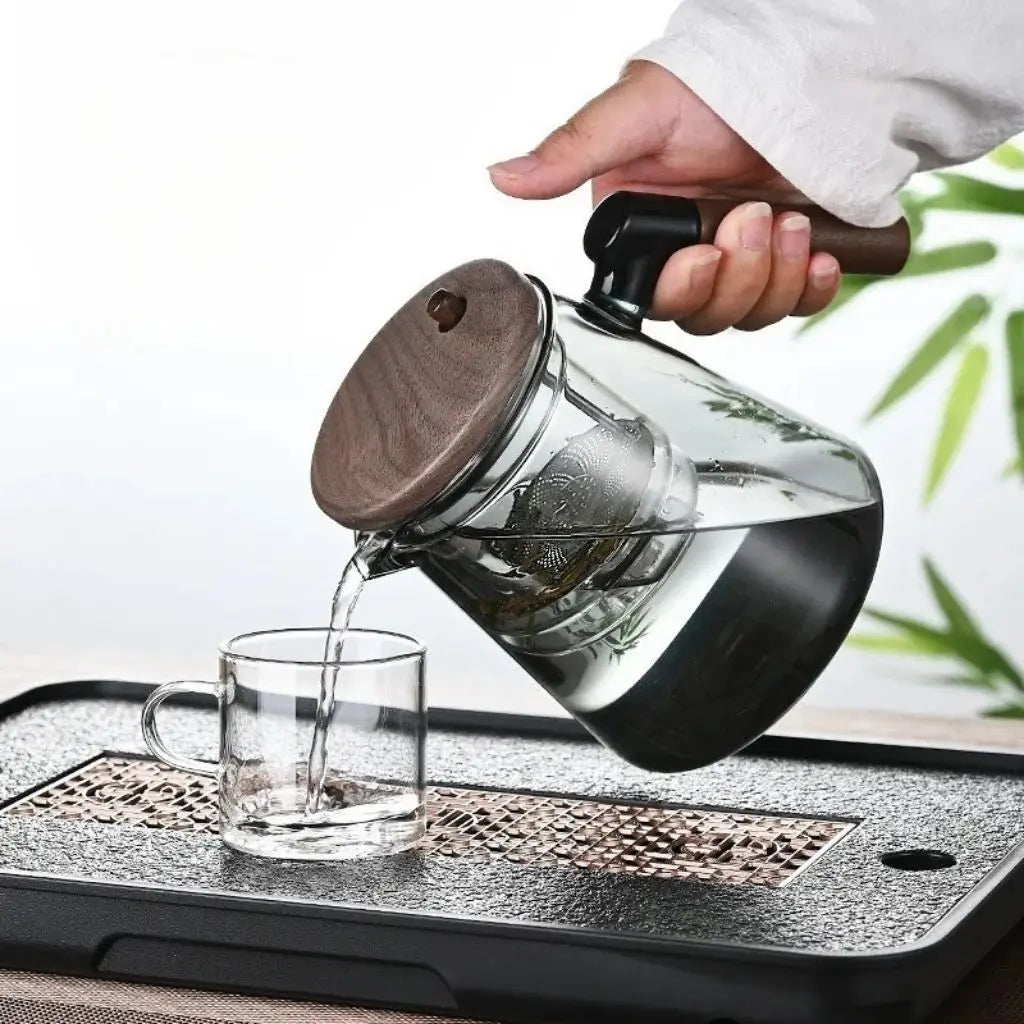 Glass teapot with wooden lid being filled with tea into a glass mug on a tray.