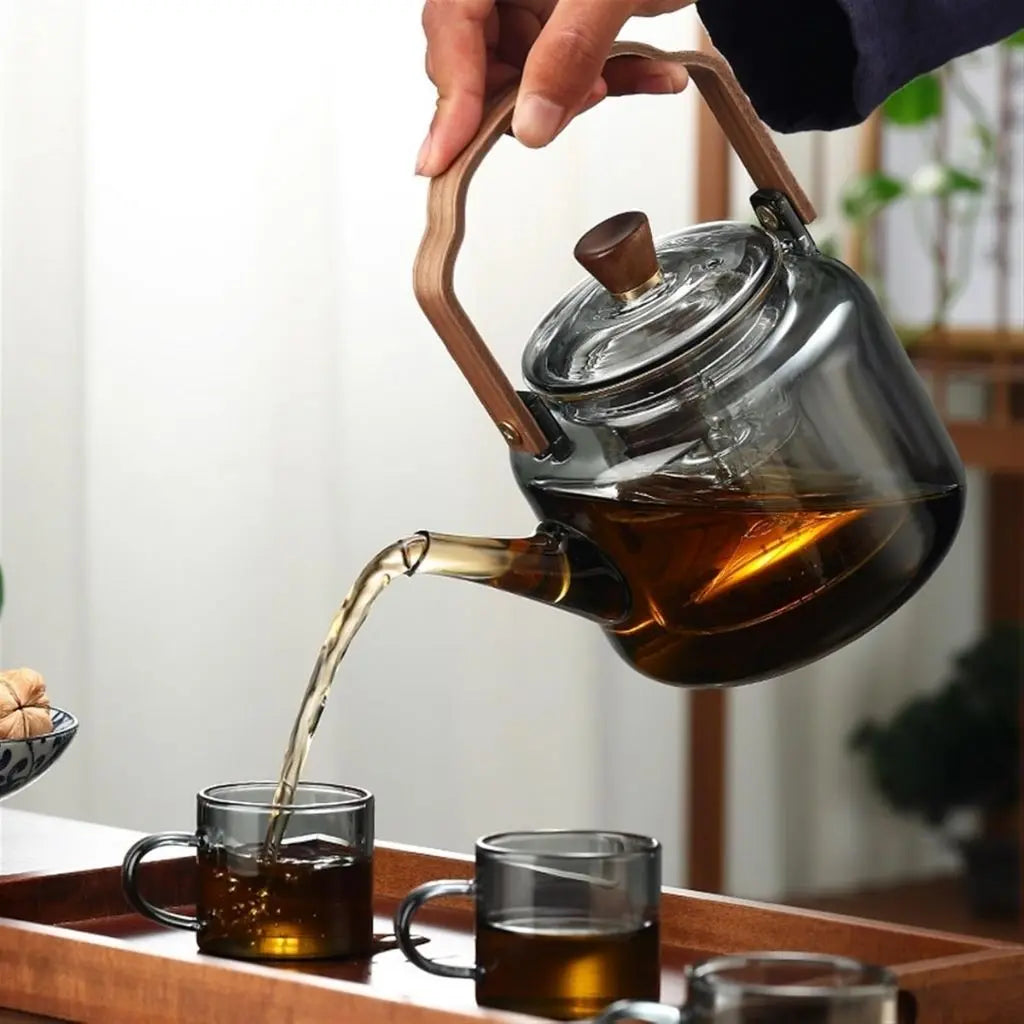 Glass teapot with wooden handle being poured into glass cups on a wooden tray.