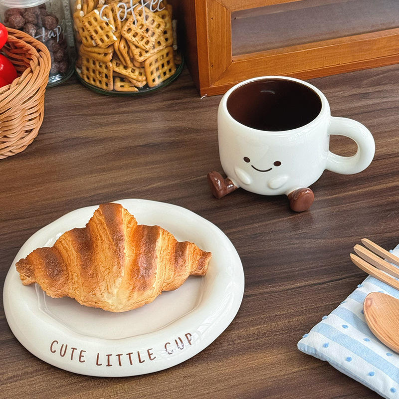Cute ceramic cup with a smiley face on a wooden surface with a croissant and a plate.