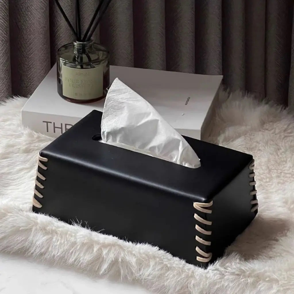 Black tissue box with white tissues on a fluffy surface with a candle and book in the background.