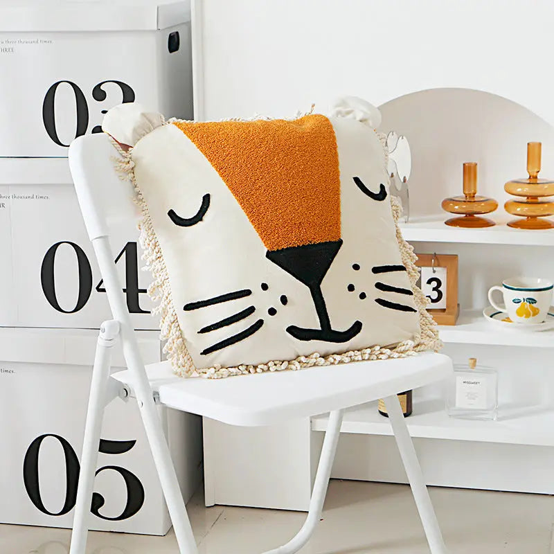 A decorative pillow with a lion face design, featuring a beige mane, black facial features, and orange ears, placed on a white chair.