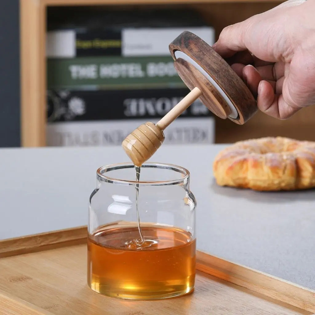Person using a wooden honey dipper to pour honey into a glass jar on a wooden tray.