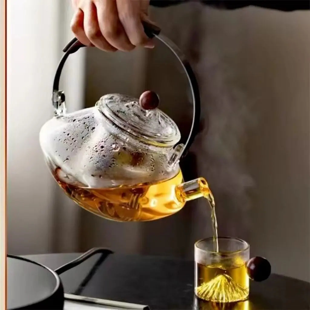 Glass teapot being filled with tea from a kettle, steam rising, on a blurred background.