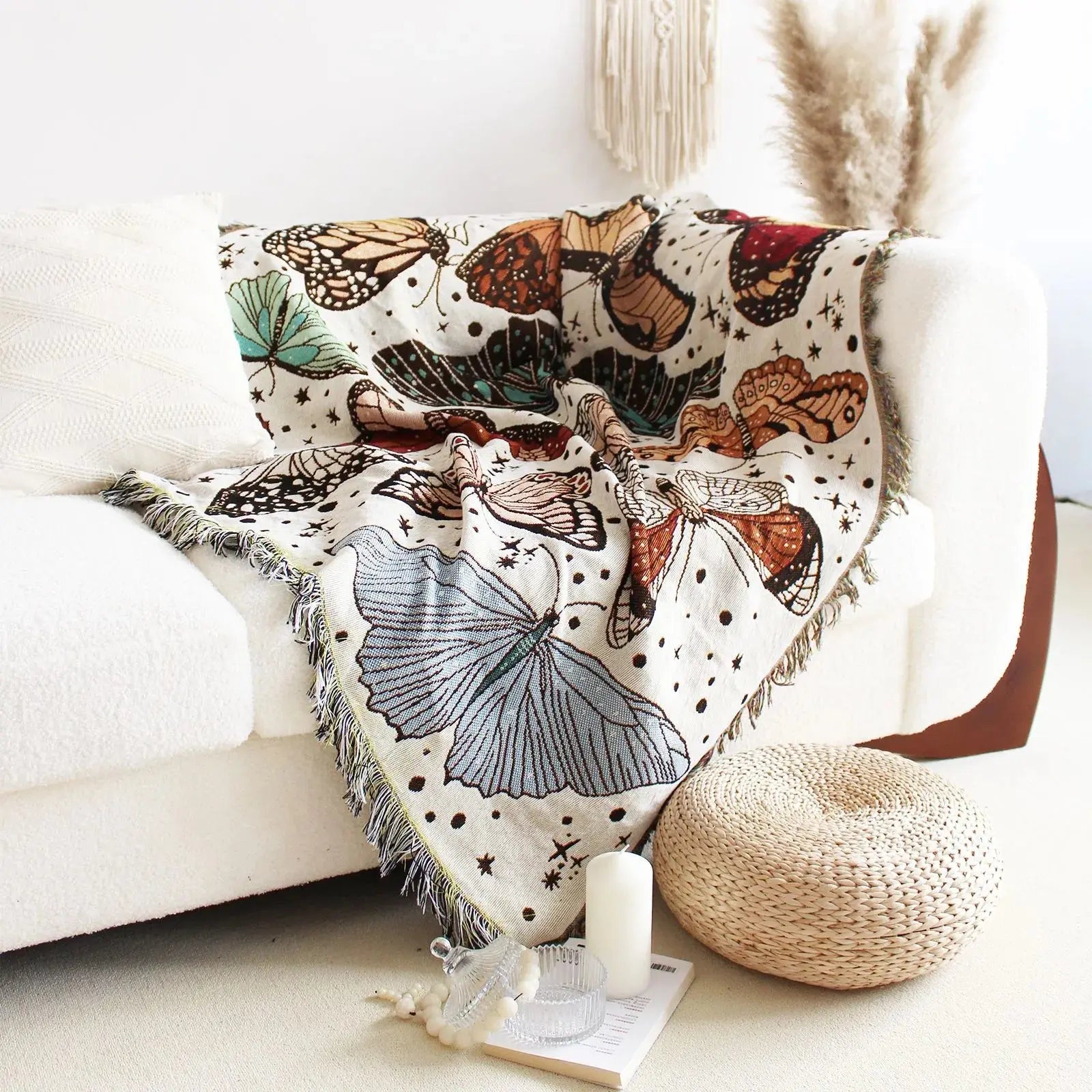Butterfly patterned blanket draped over a white couch with a woven pouf and candle in the foreground.