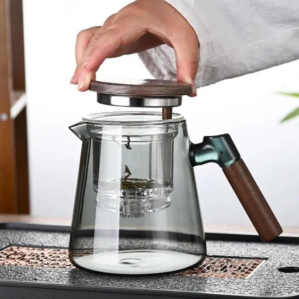 Glass teapot with wooden handle and lid being held by a hand on a textured surface.