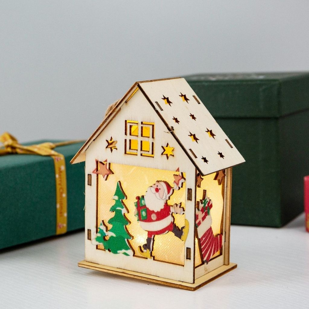 Decorative house-shaped lantern with Santa Claus and Christmas tree design, placed on a surface with gift boxes in the background.