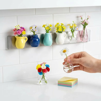 Colorful vases with flowers on a wall-mounted holder, with a hand holding a small clear vase.